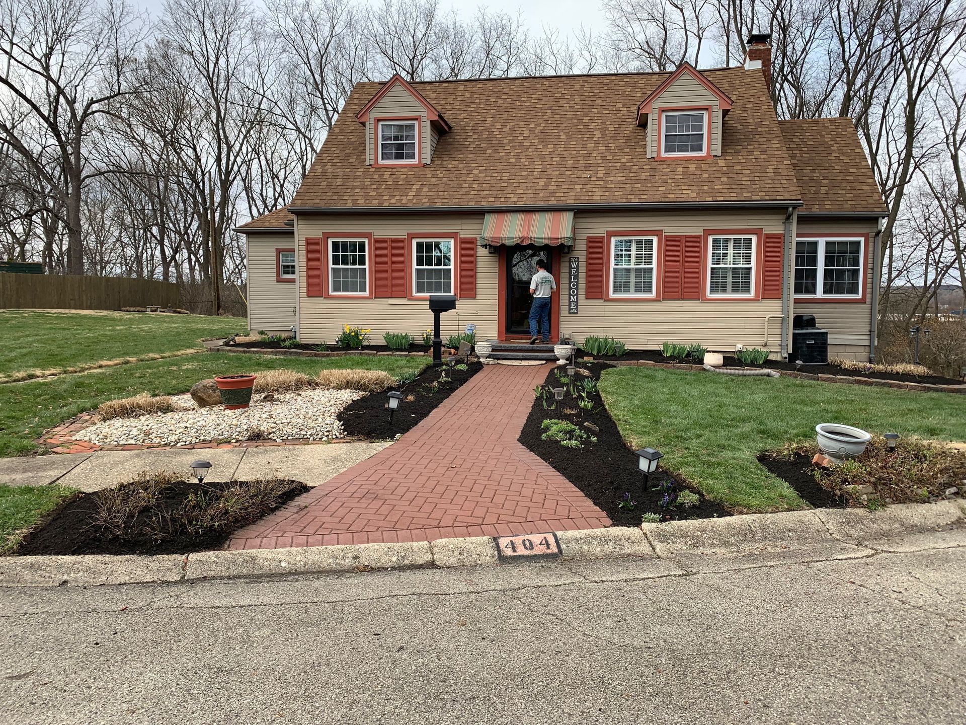 Tan house with brick walkway and red shutters, person stands in doorway.