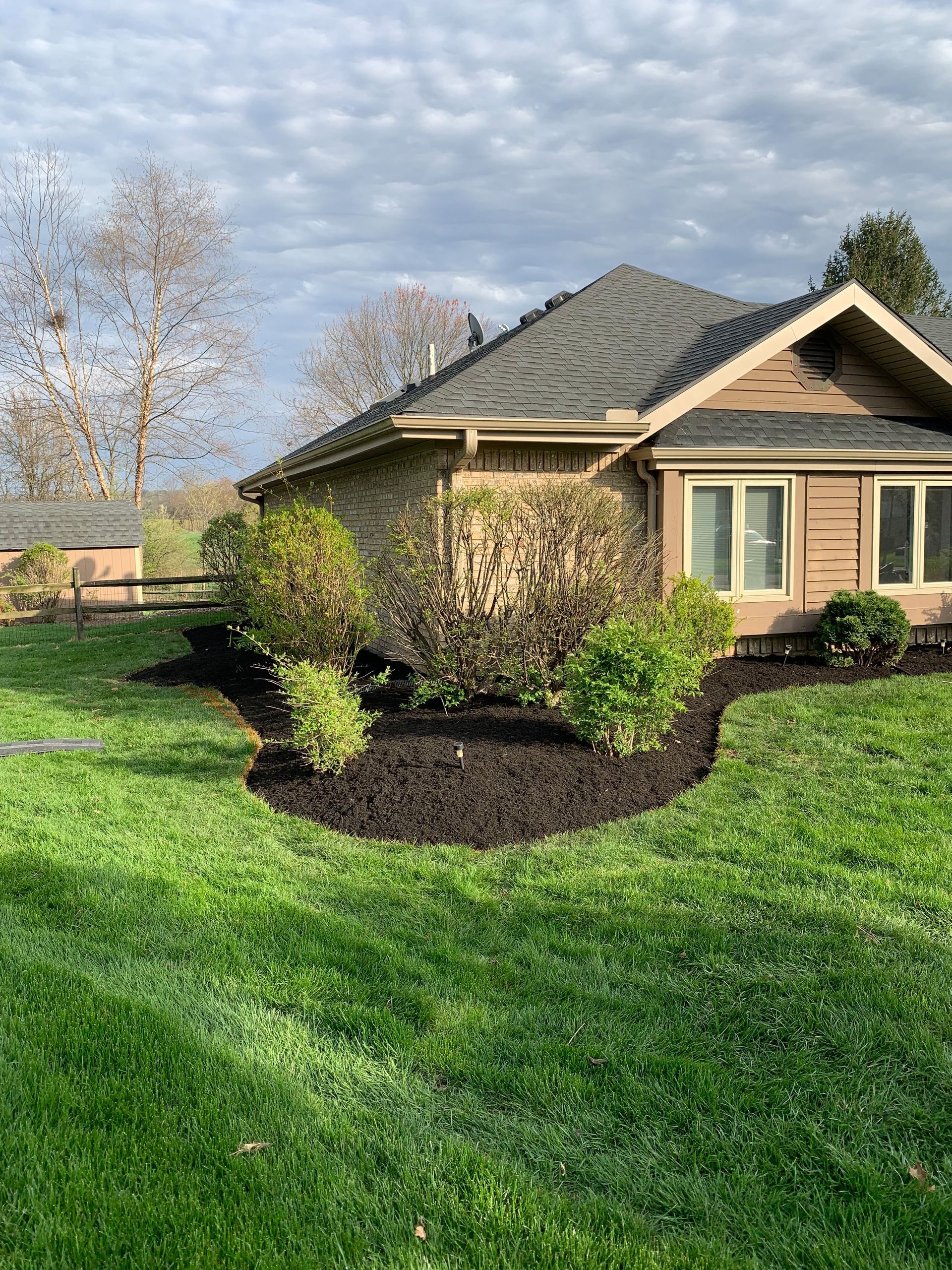 A tan house with brown shingles and trim; dark mulch beds around shrubs, on green grass under a cloudy sky.