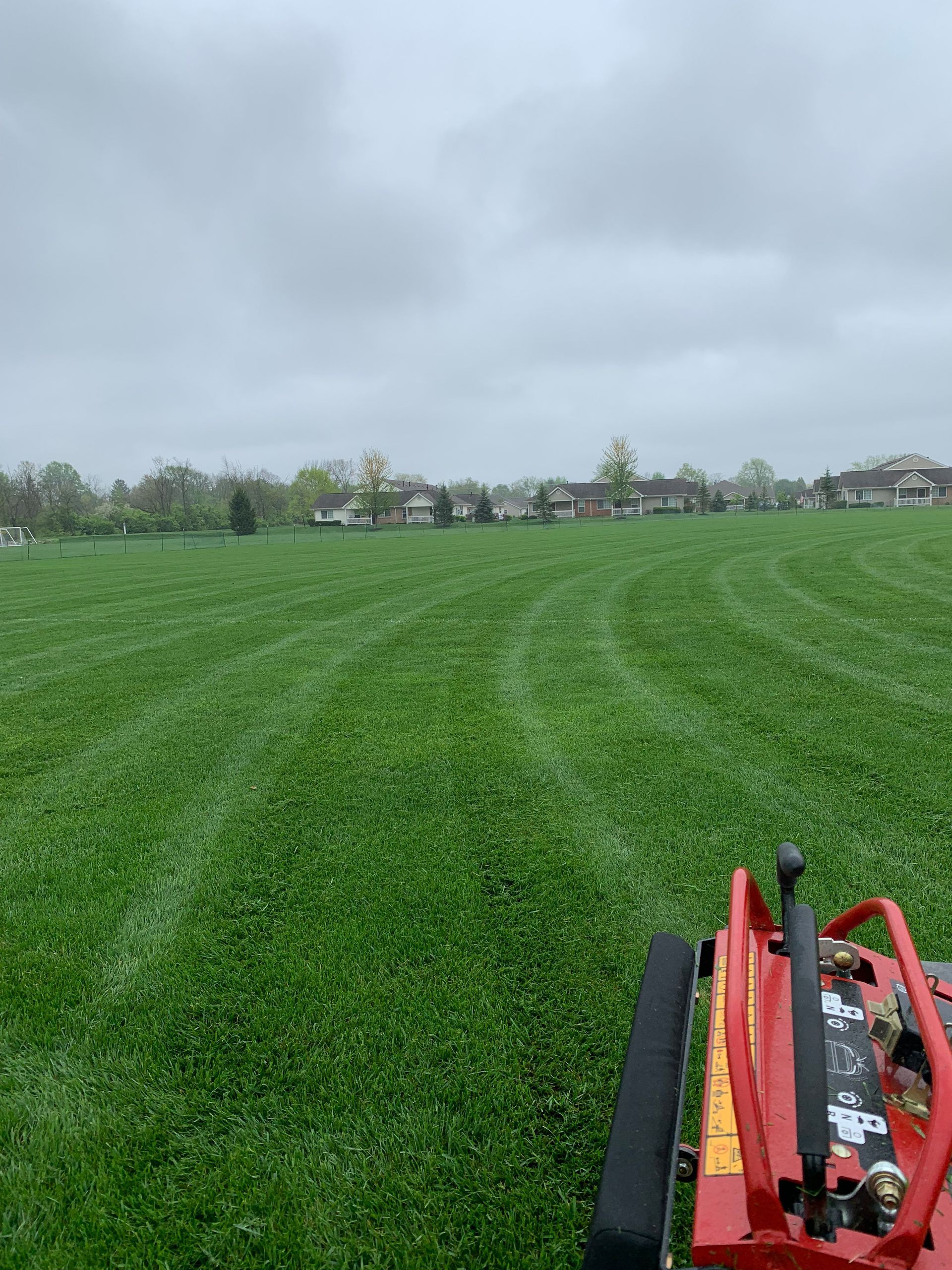 Lawnmower cutting stripes in a large green field, overcast sky, houses in the distance.