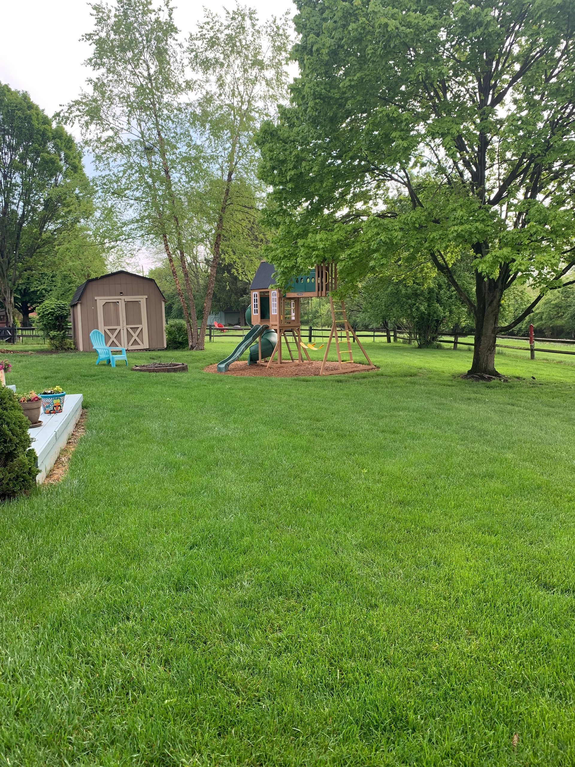 Lush green backyard with playset, shed, and trees. Overcast sky, open space, and small house in background.
