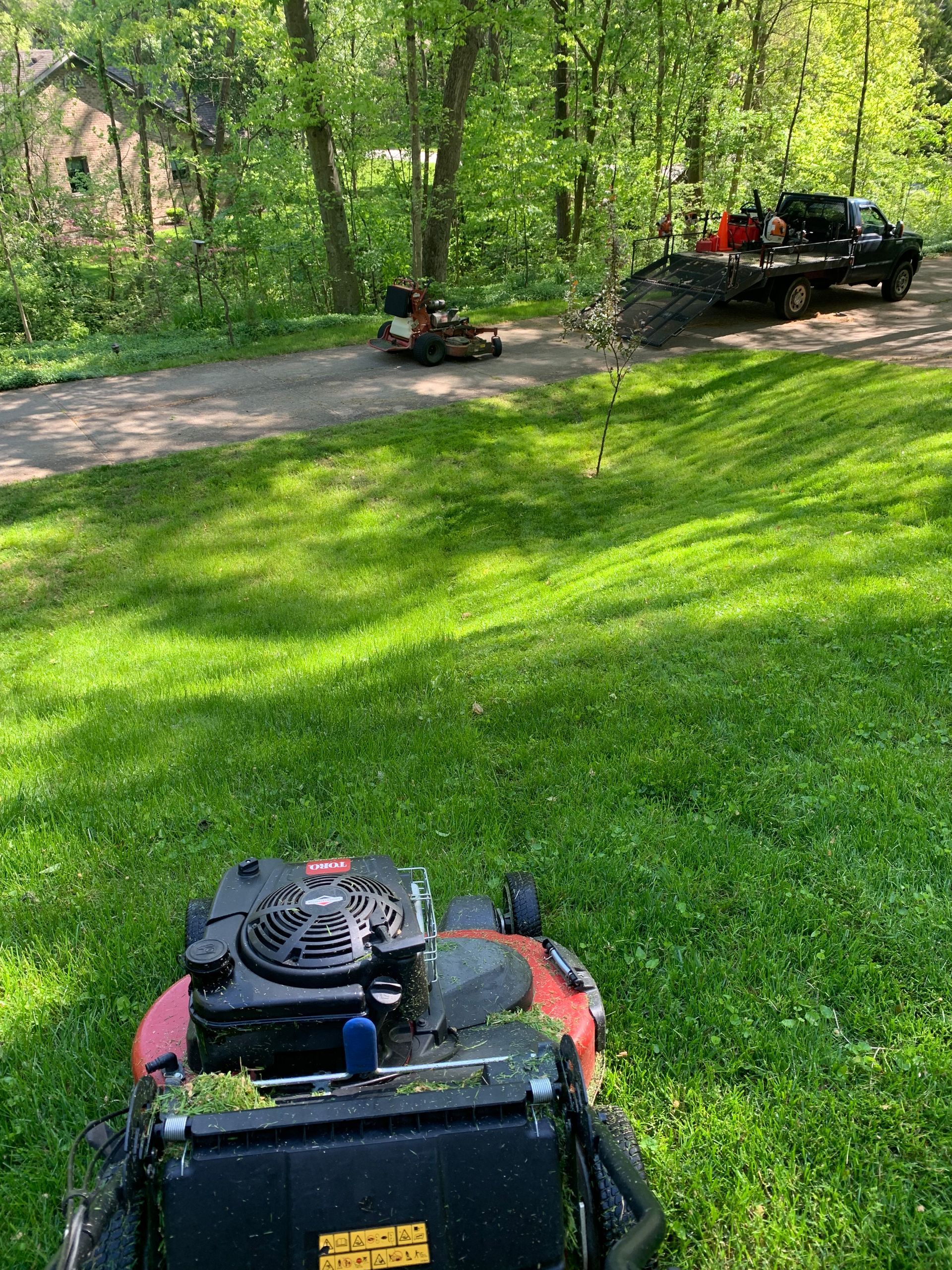 A person mowing a grassy area with a lawnmower; another mower and truck in the background.
