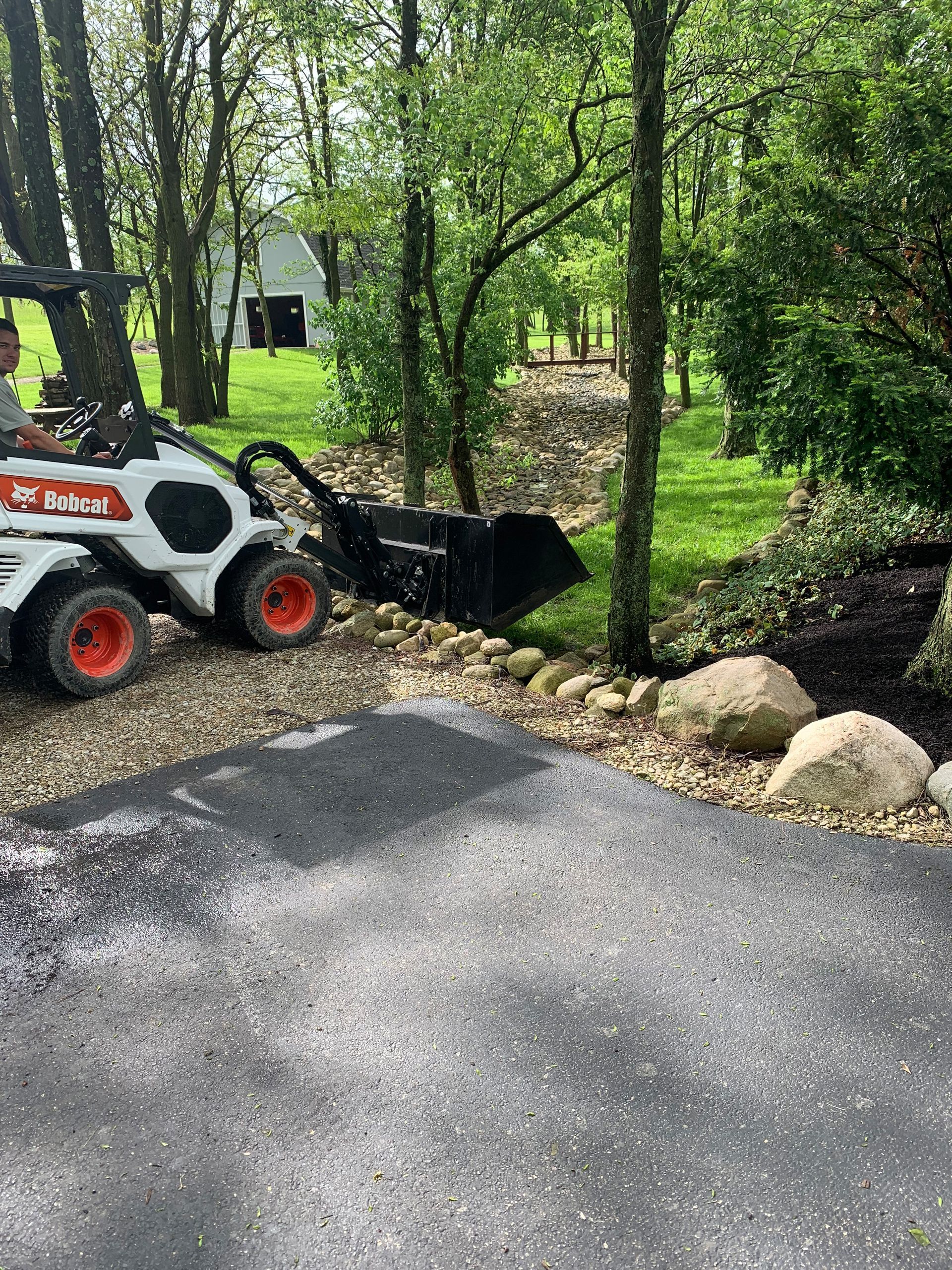 A man operates a Bobcat, moving gravel in a driveway. Green trees and grass surround the area.