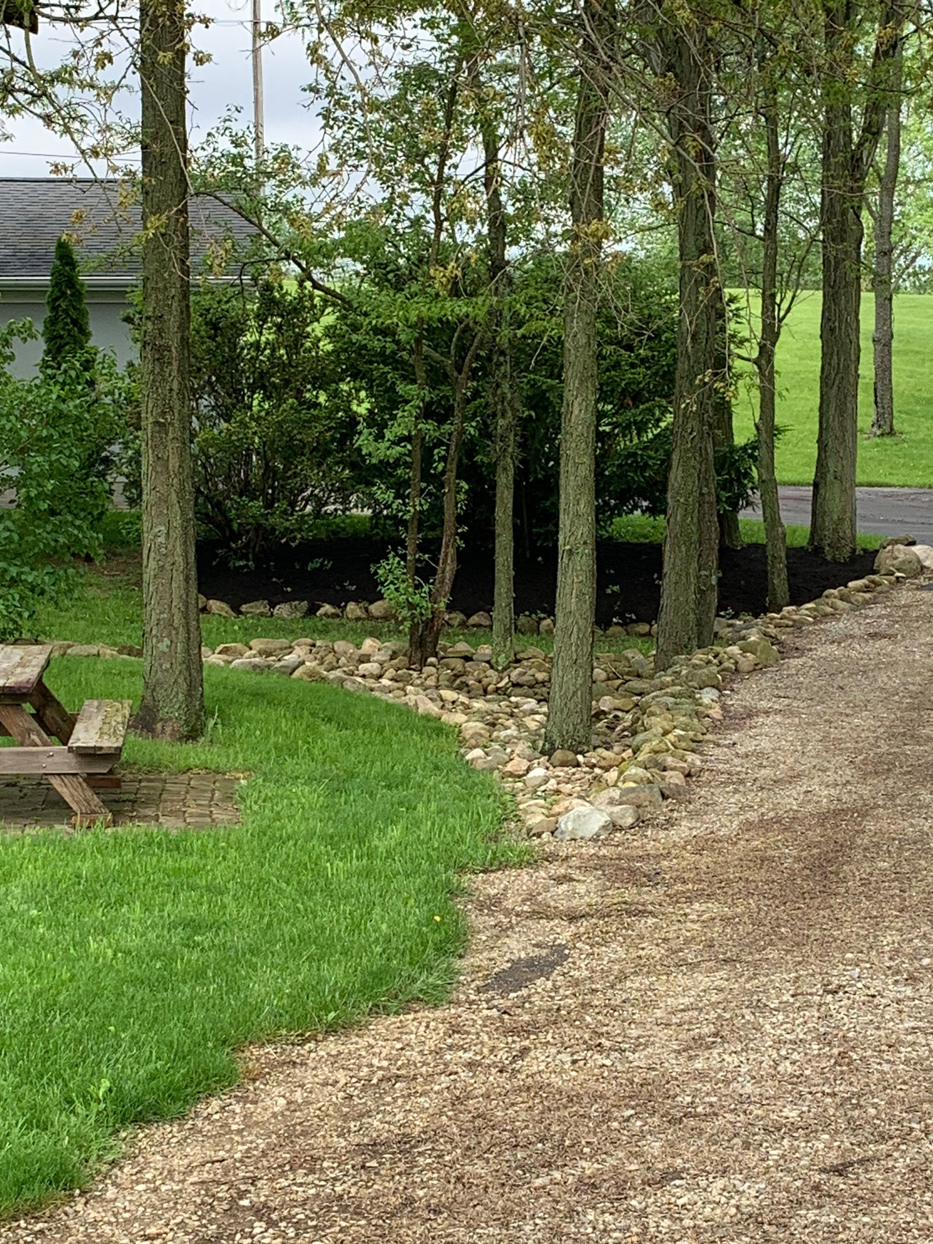 Gravel driveway curves past trees and a grassy lawn with a picnic table. A building is in the background.