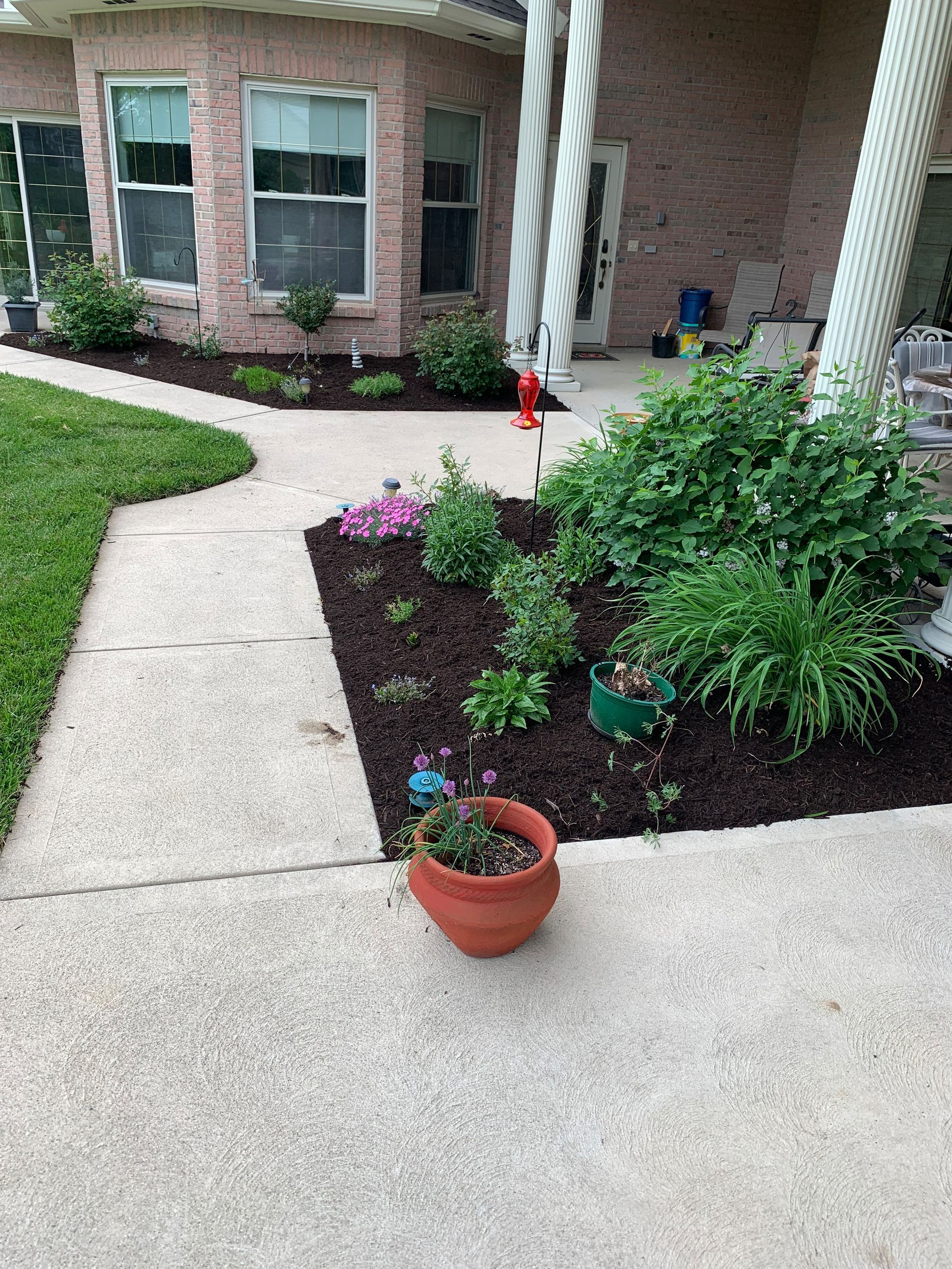 A cement path leads past a landscaped flower bed with a brick house in the background.