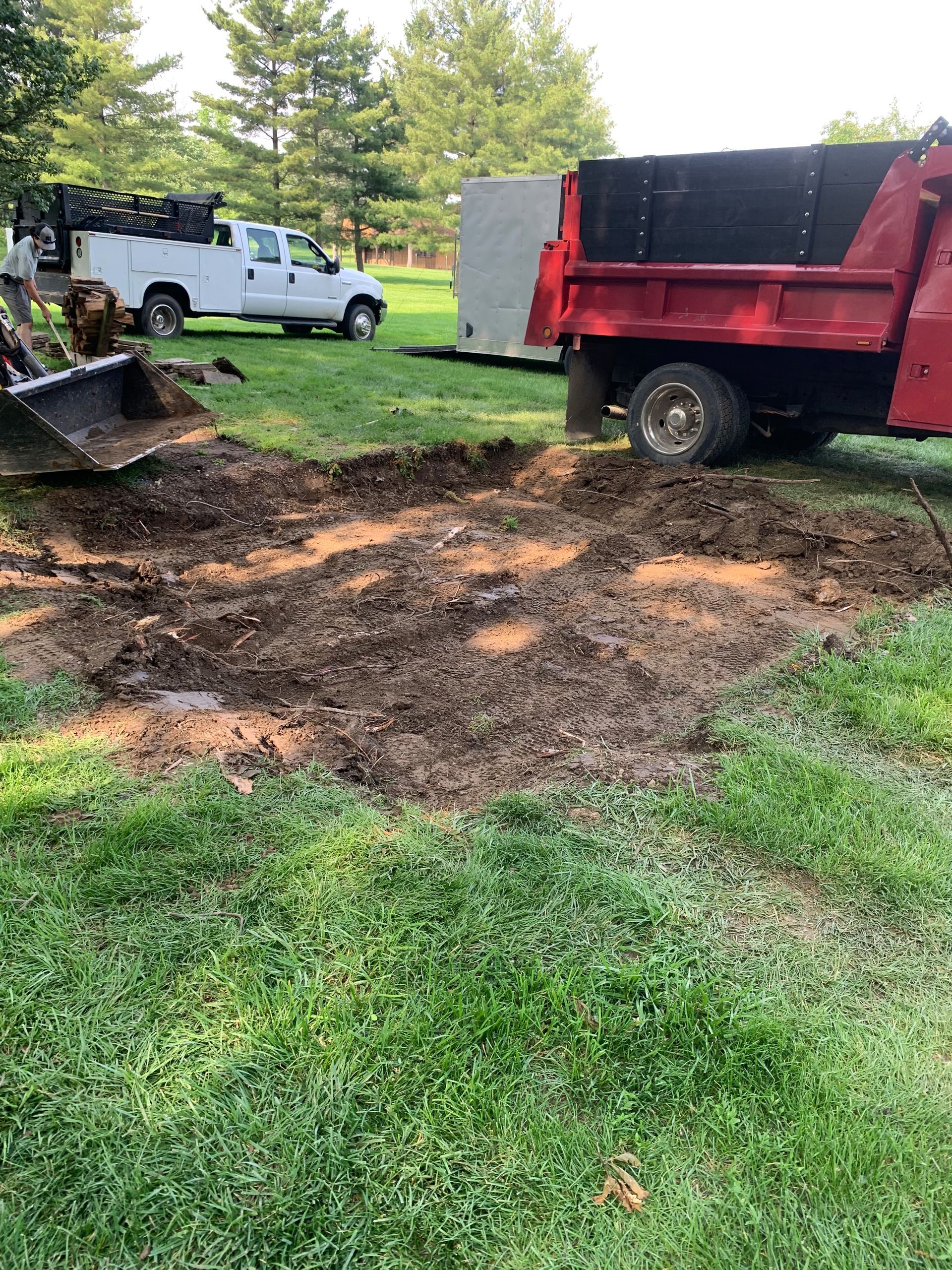 A red dump truck and white truck with a backhoe are removing dirt from a grassy area.