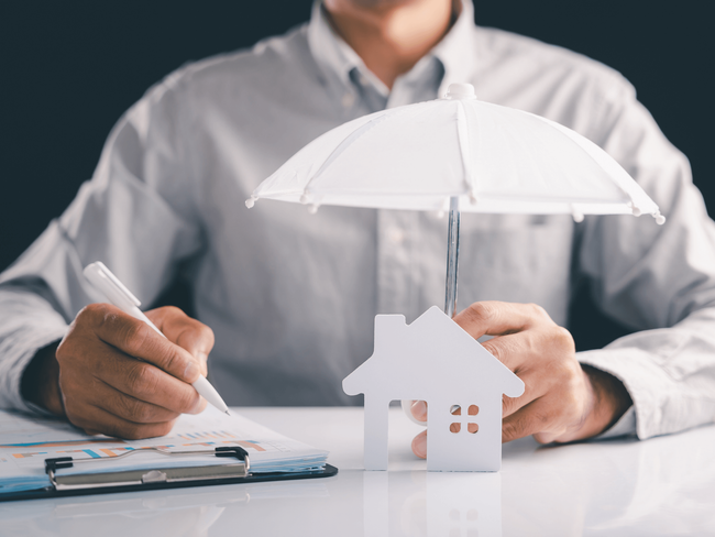 Person holding an umbrella over a small house model while signing paperwork, representing protection from a home and auto insurance broker service in Maricopa, AZ