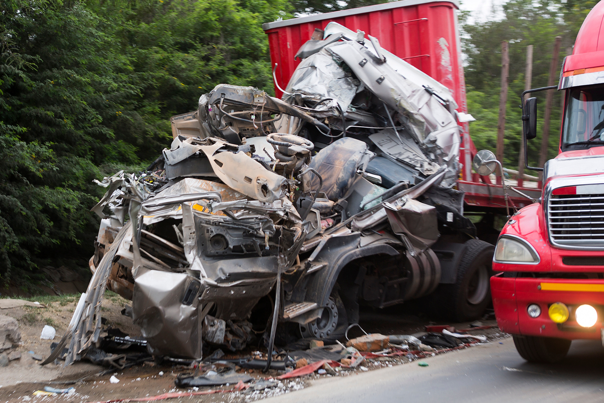 A red semi truck is driving past a wrecked truck on the side of the road.