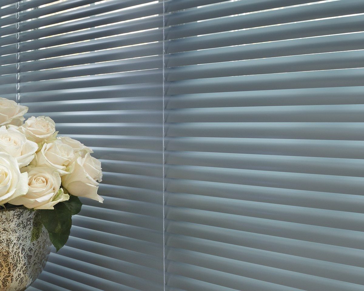 A vase of white roses sits in front of a window with blinds