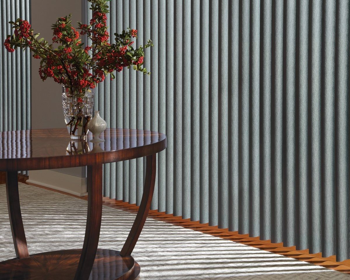 A table with a vase of flowers on it in front of vertical blinds.