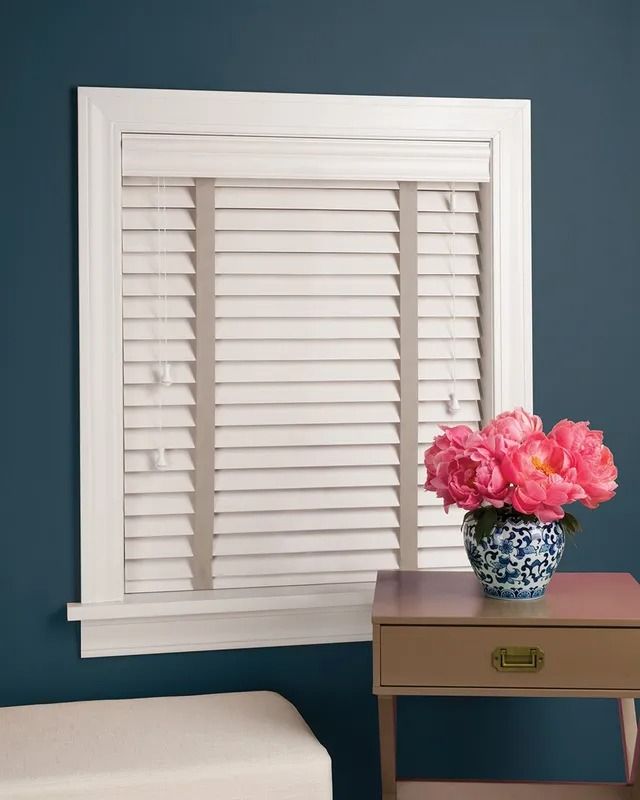A window with white blinds and a vase of pink flowers on a table