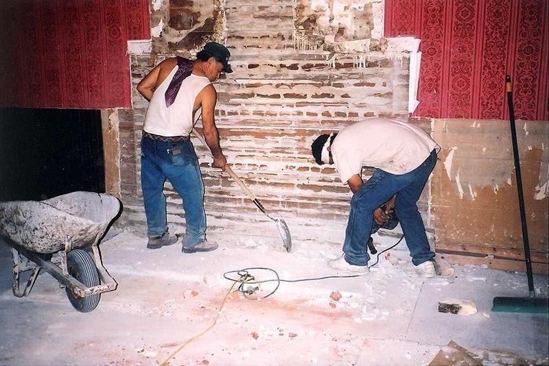 Two men are working on a brick wall with a wheelbarrow in the foreground