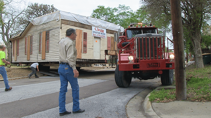 A red truck is carrying a house down a street