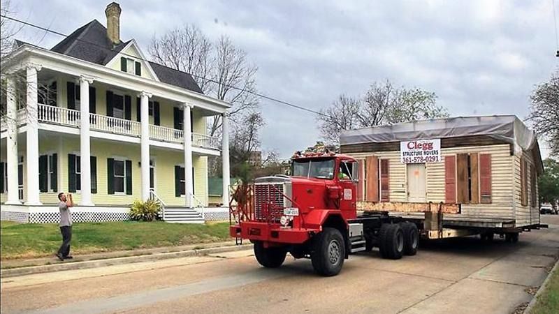 A red truck is carrying a house down a dirt road.