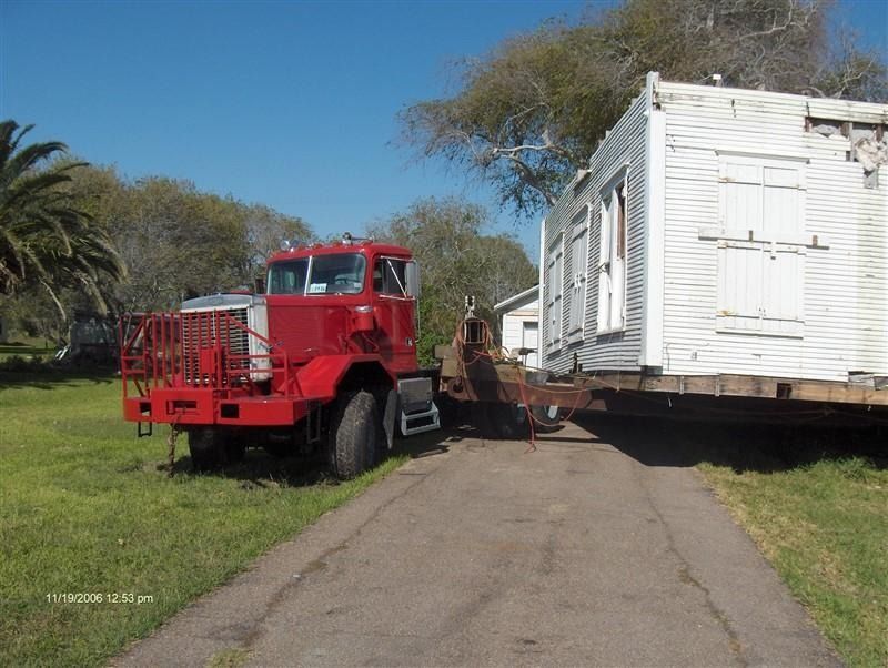 A red truck is carrying a white house down a road