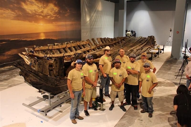 A group of men are standing in front of a large wooden boat.