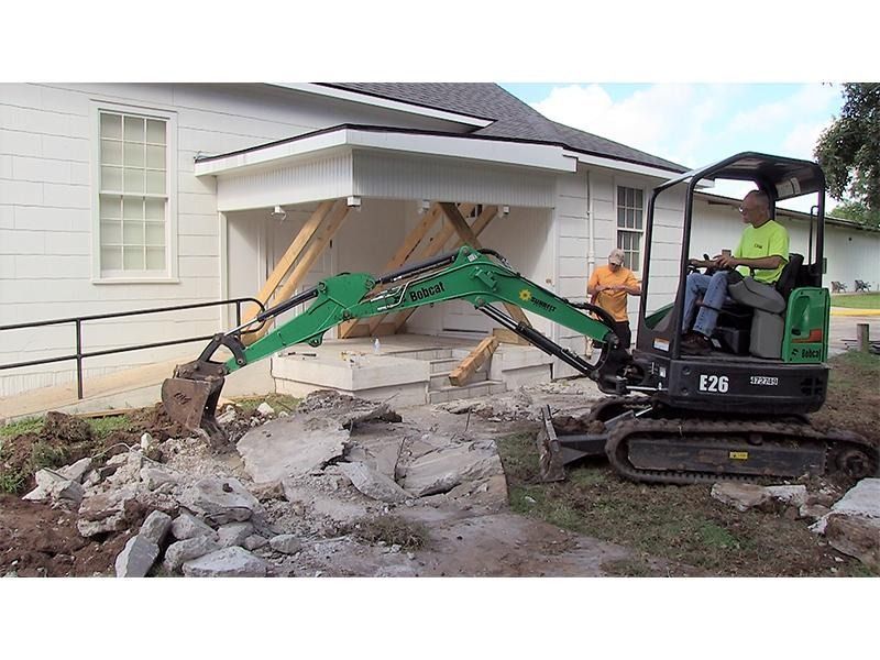 A man is driving a green excavator in front of a house.