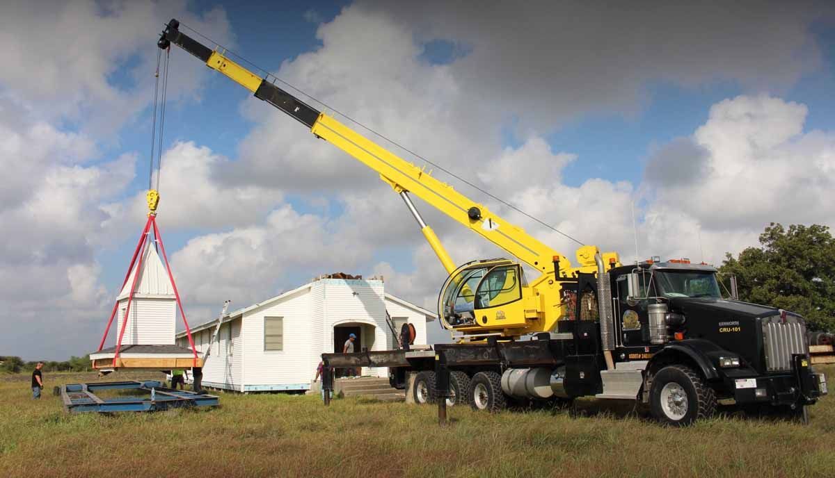 A yellow crane is lifting a piece of wood in front of a house.