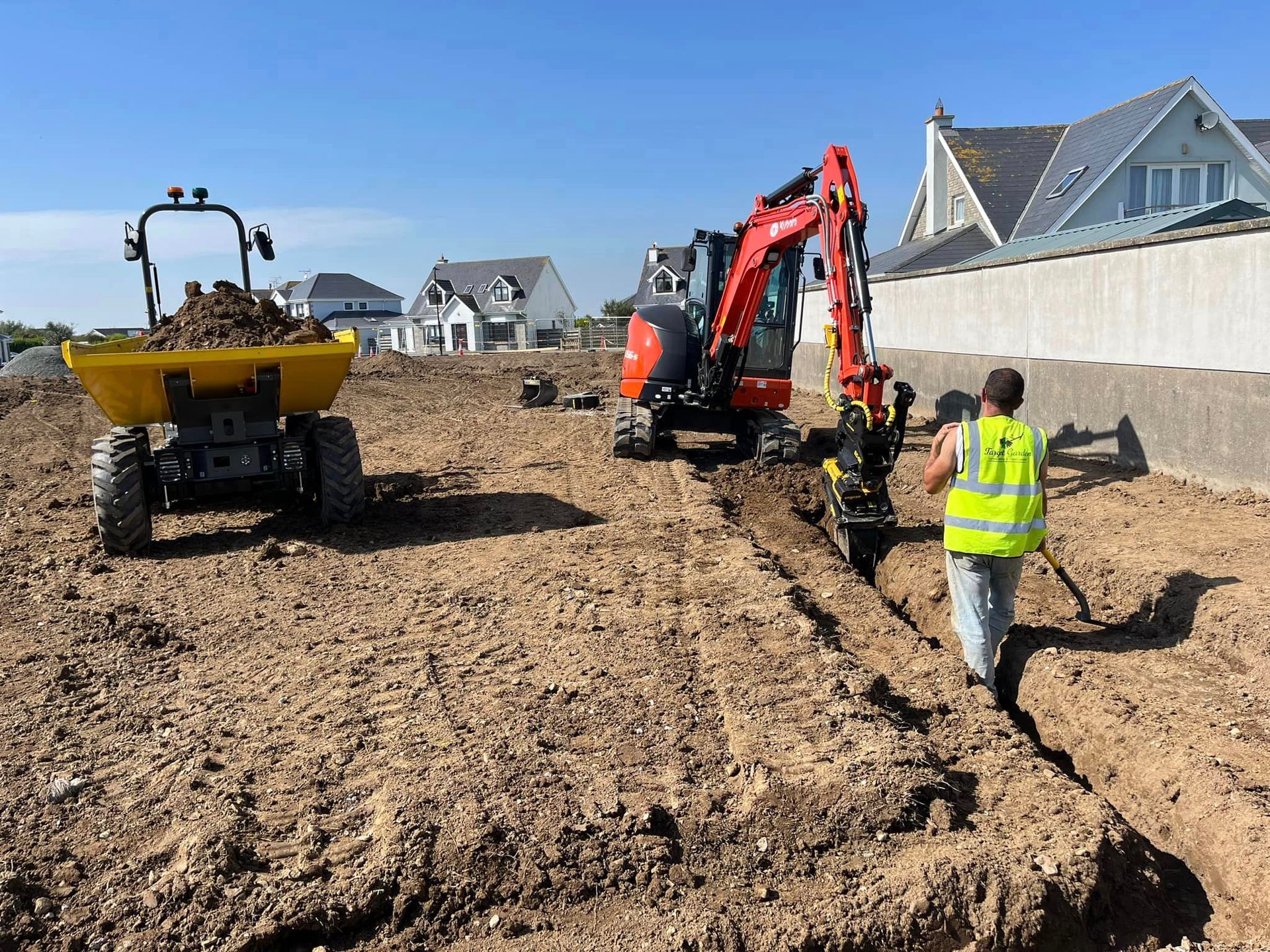 A man is walking in the dirt next to a dump truck and an excavator.