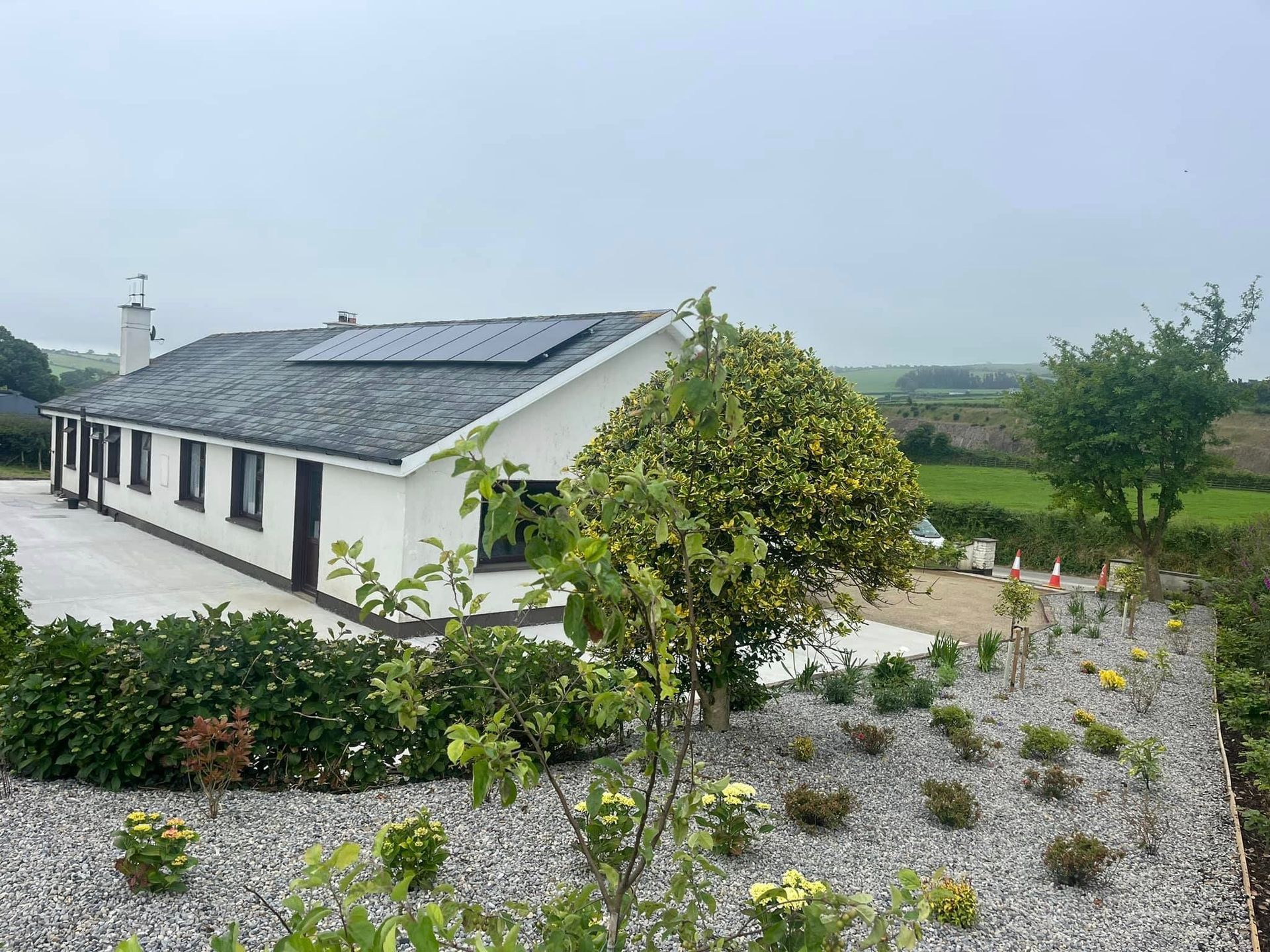 A white house with solar panels on the roof is surrounded by gravel and trees.