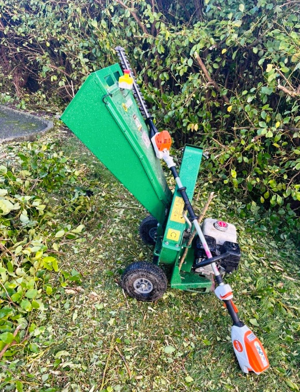 A green tree chipper is sitting in the grass next to a hedge trimmer.