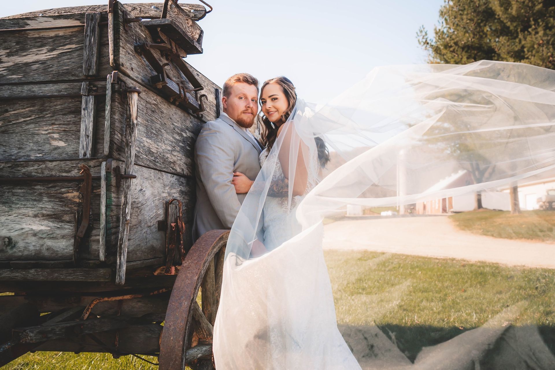 A bride and groom are posing for a picture in front of an old wooden wagon.