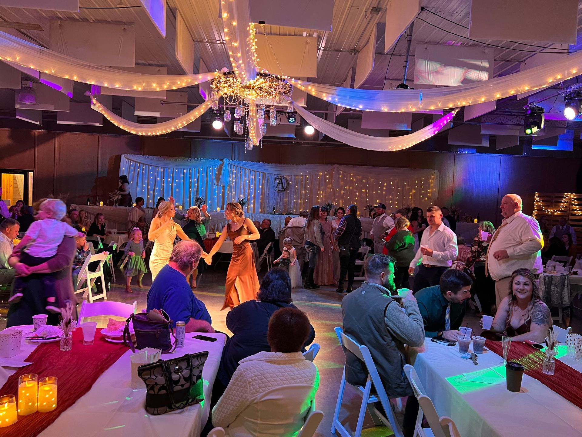 A group of people are dancing in a large room at a wedding reception.