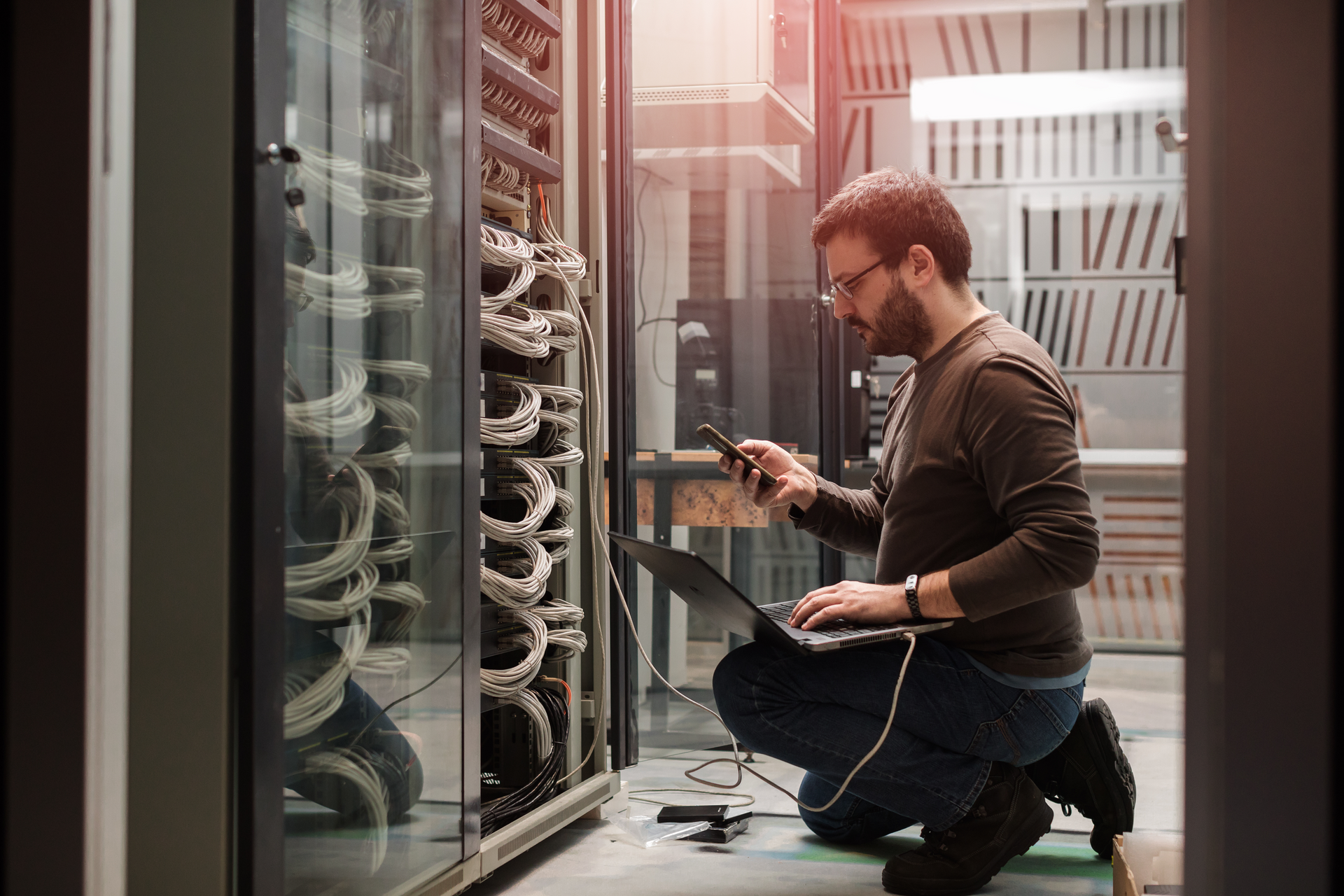 A man is kneeling down in front of a server with a laptop.