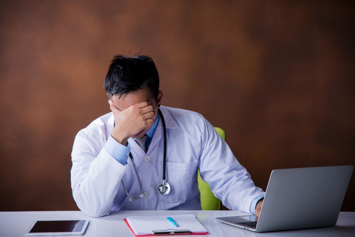 A doctor is sitting at a desk with a laptop and covering his face.