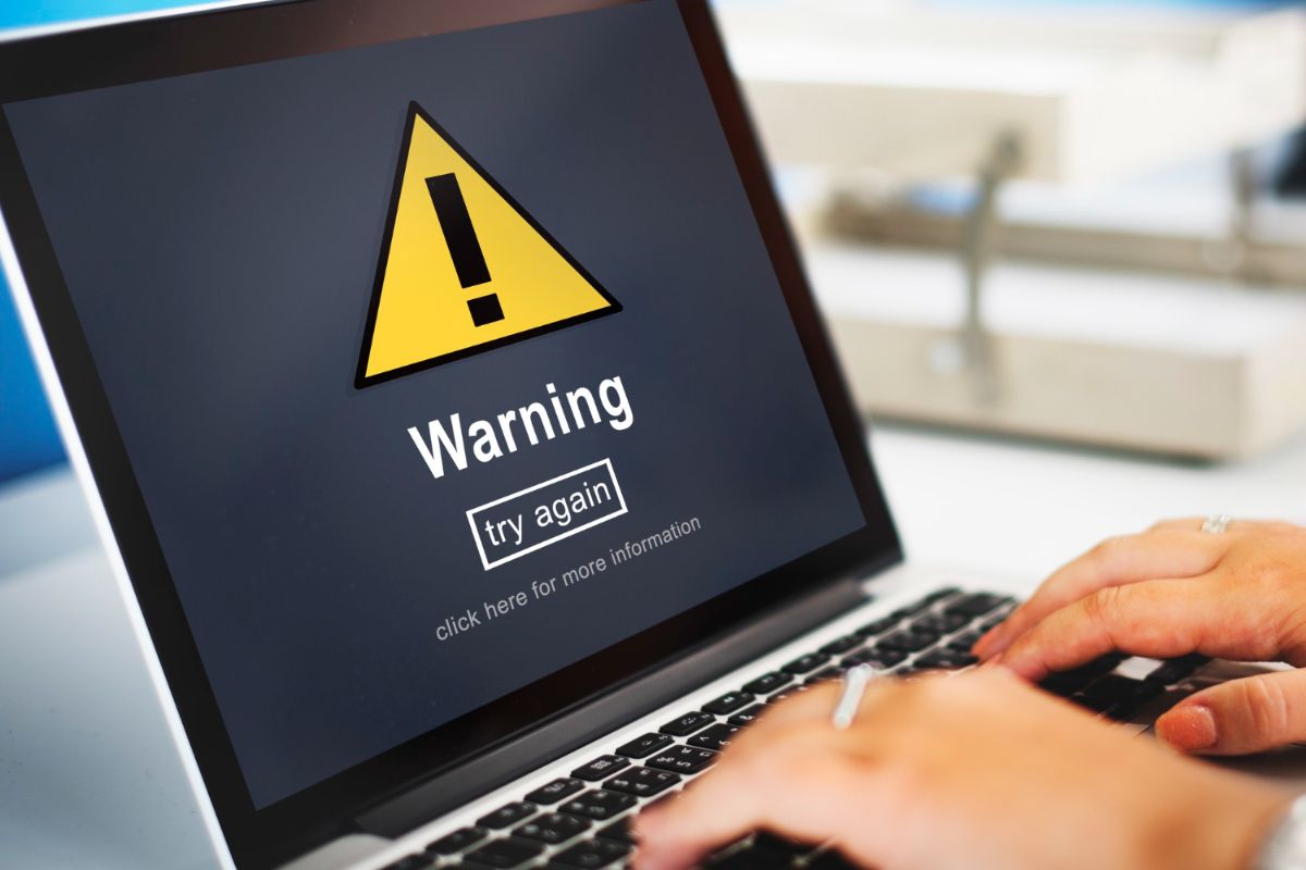 A person is typing on a laptop with a warning sign on the screen.