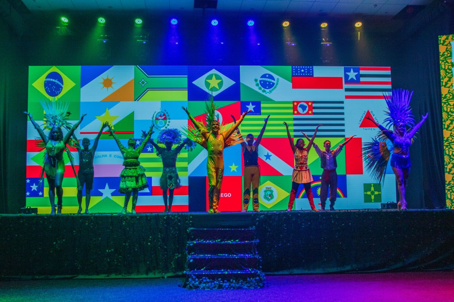 Um grupo de pessoas está em um palco em frente a uma grande tela com bandeiras.