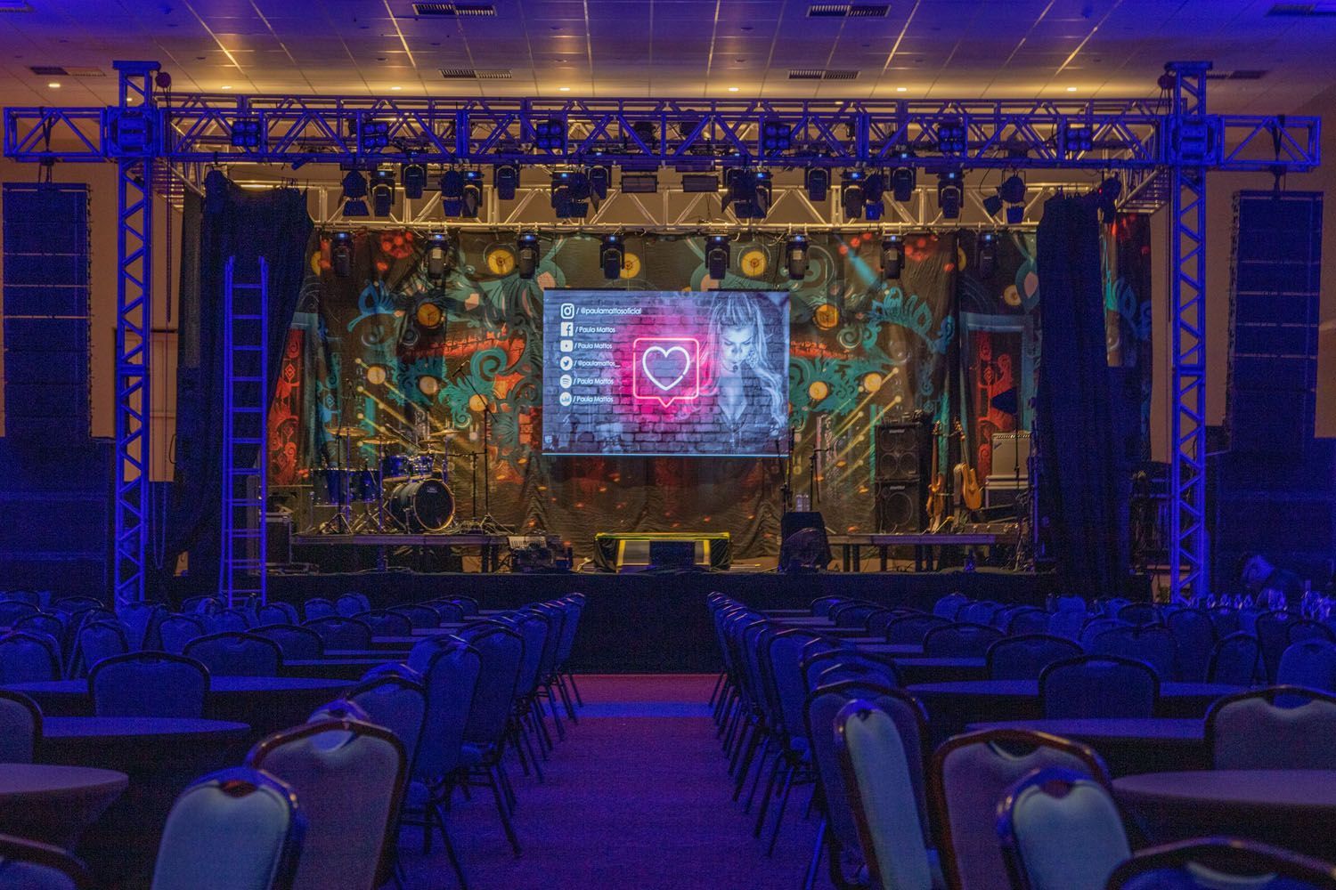 Um auditório vazio com mesas e cadeiras e um palco com telão.