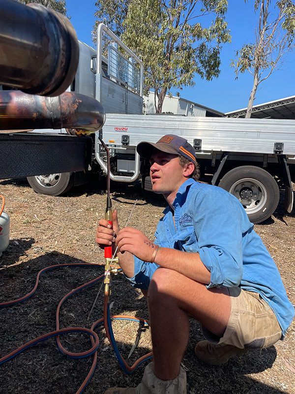 A Man Is Kneeling in Front of A Truck While Welding a Pipe — BJB Plumbing & Gasfitting in Cobar, NSW