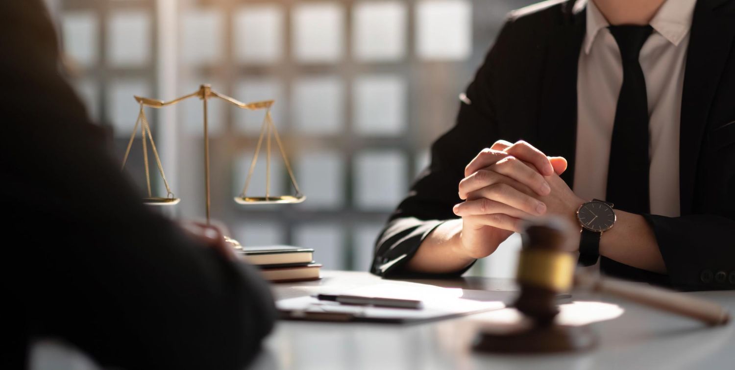 Two people in suits at a desk with a gavel, law scales, and documents.