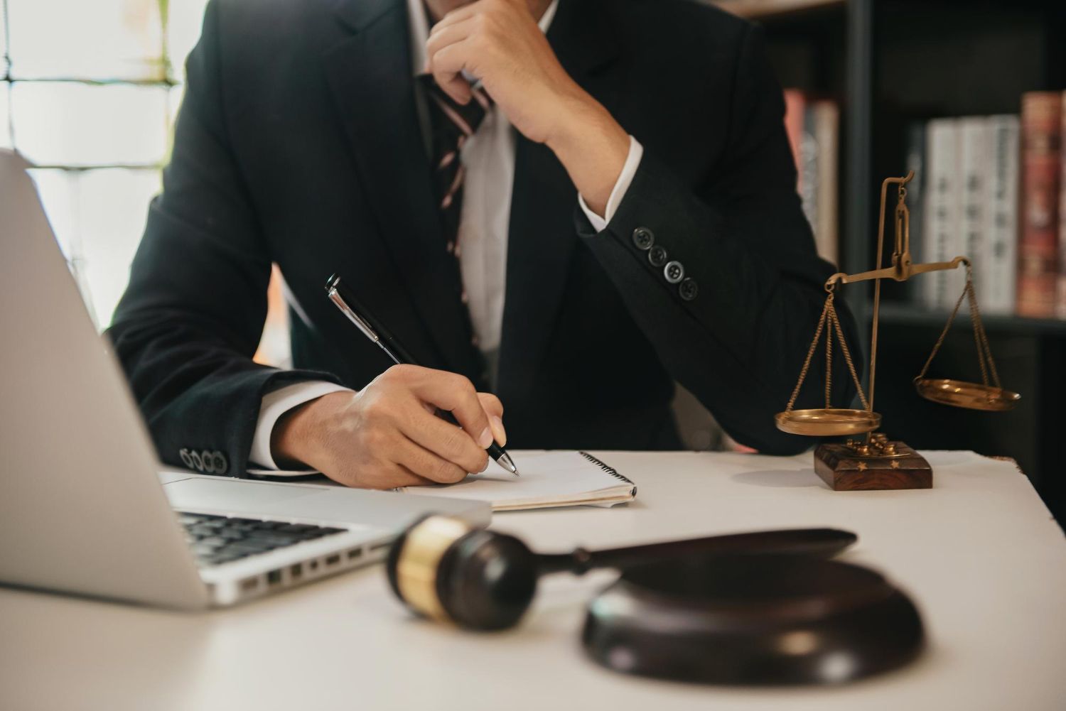 Person in suit writing, with laptop, gavel, and scales of justice on desk.