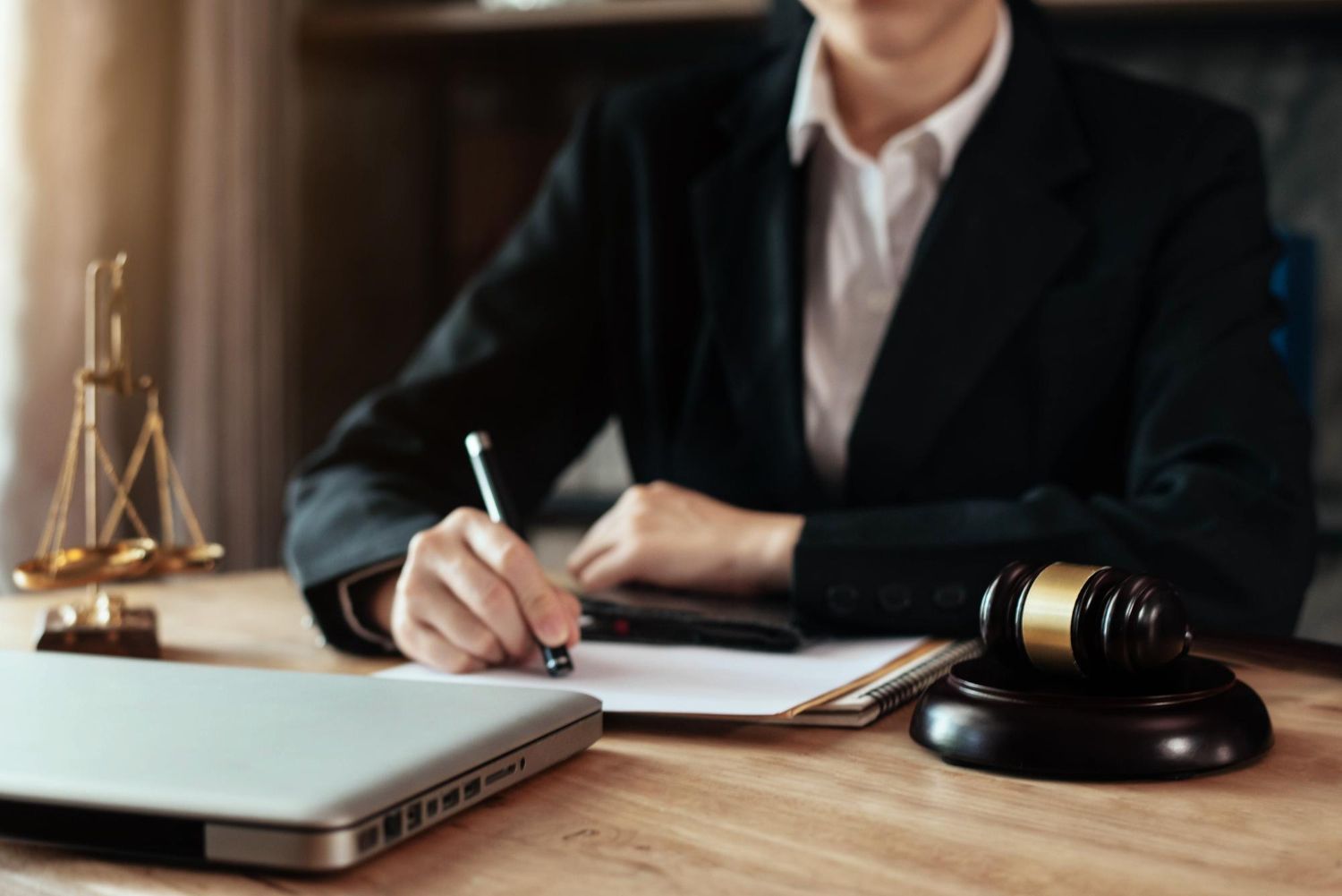Person in suit writing at a desk with a laptop, scales of justice, and a gavel.