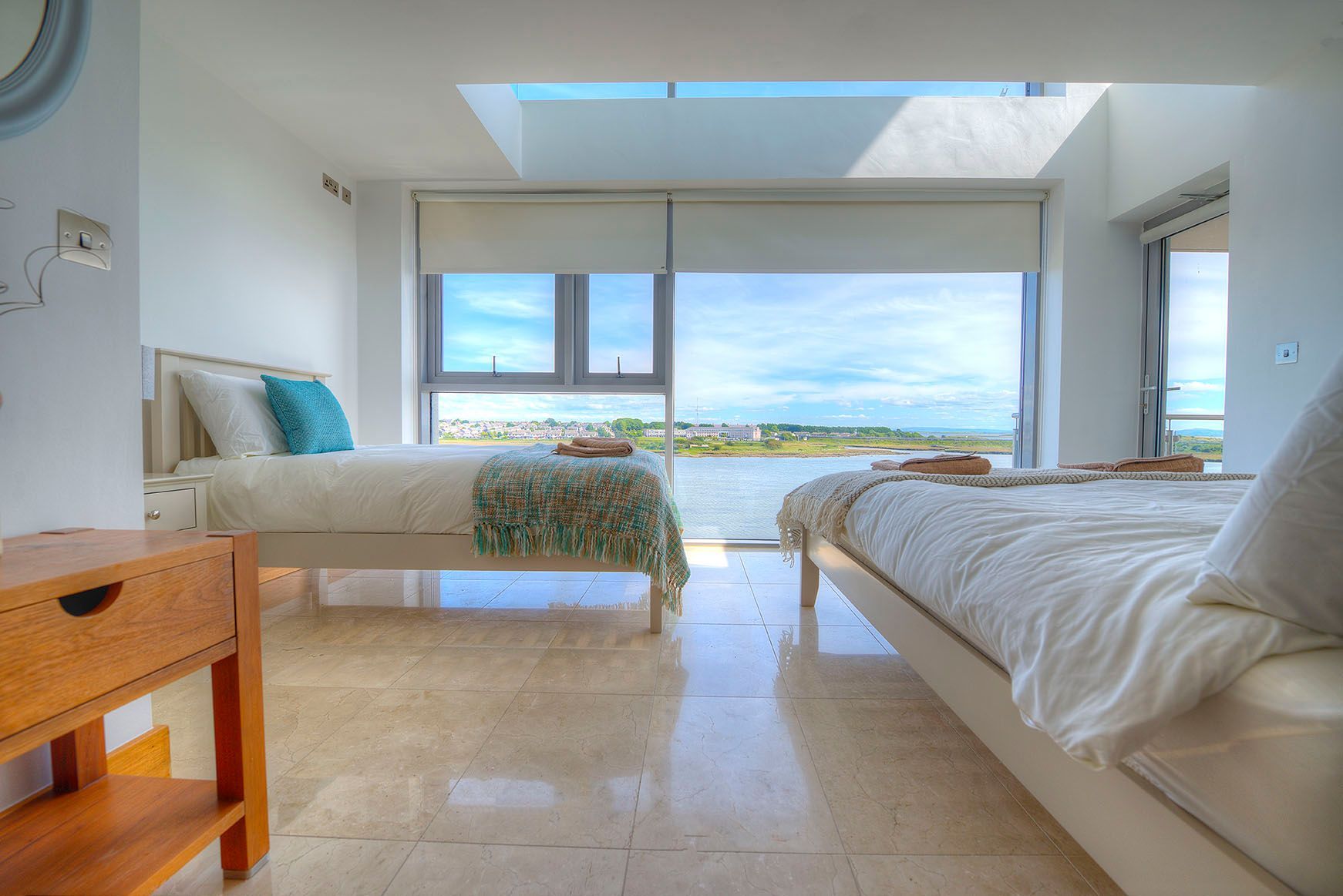 Bright bedroom with two beds, large windows overlooking a waterfront, and a small wooden nightstand.