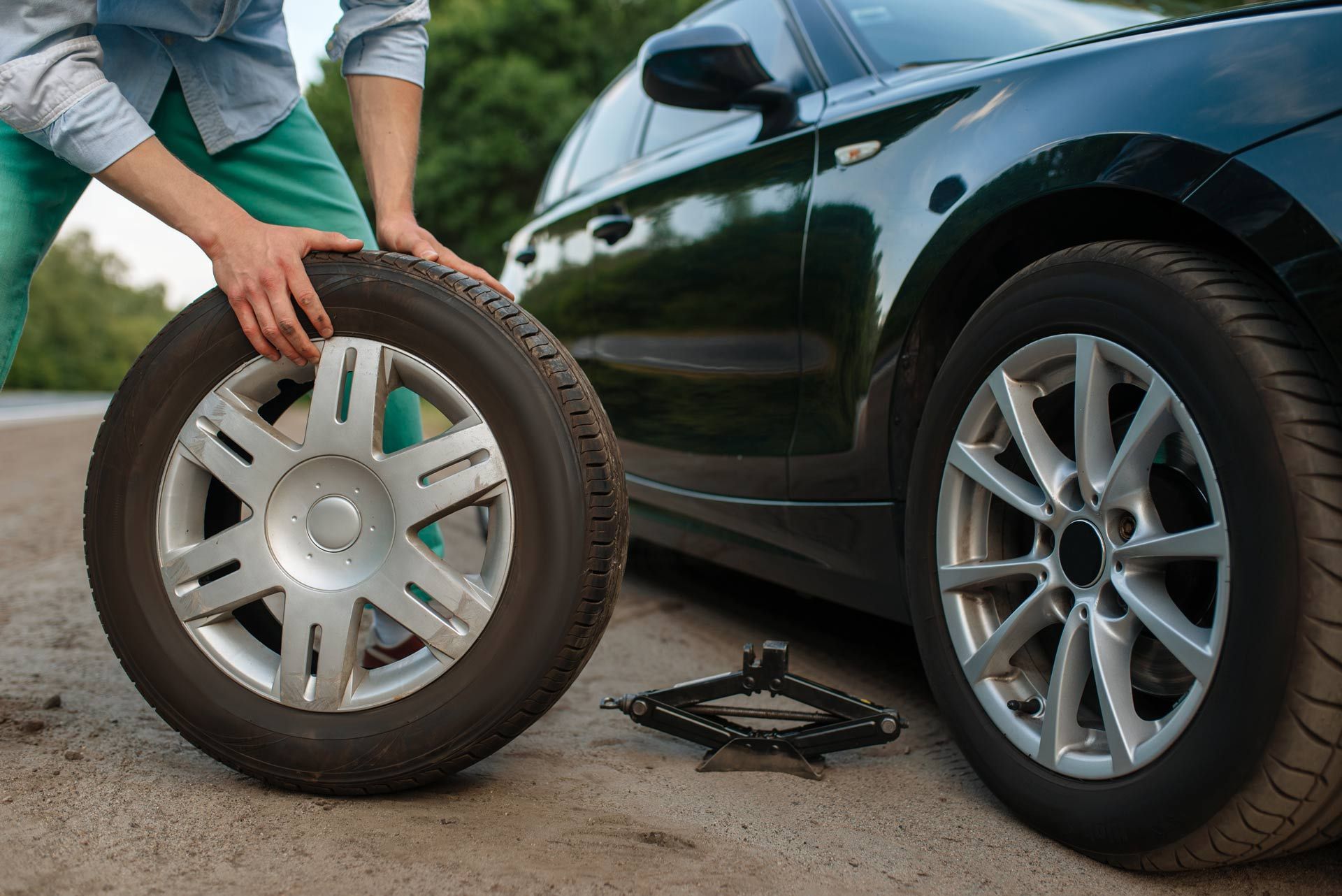 Person changing a tire beside a black car, holding a wheel with a jack on the ground.