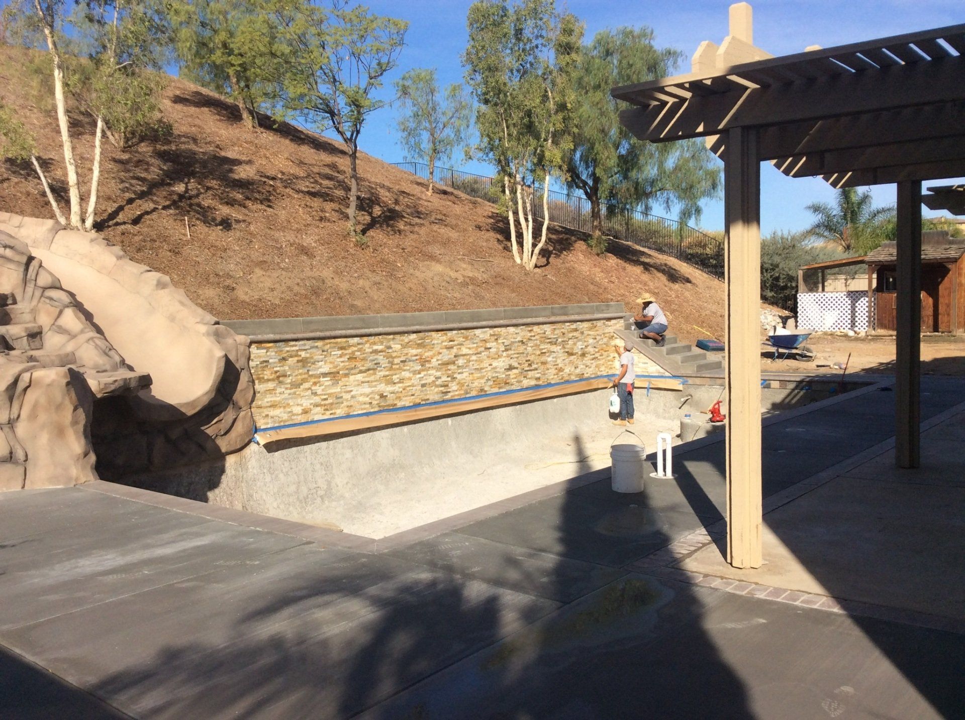 A person is sitting under a pergola next to a stone wall.