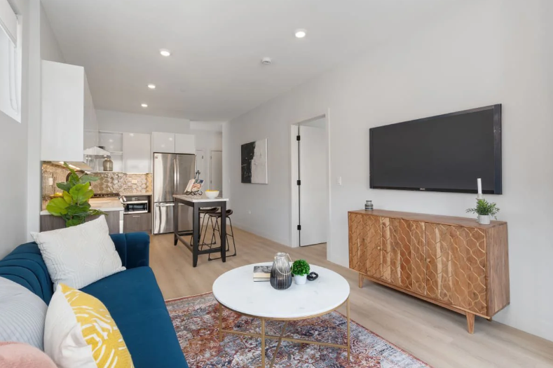 Bright modern living room with blue sofa, white coffee table, wall-mounted TV, and open kitchen area