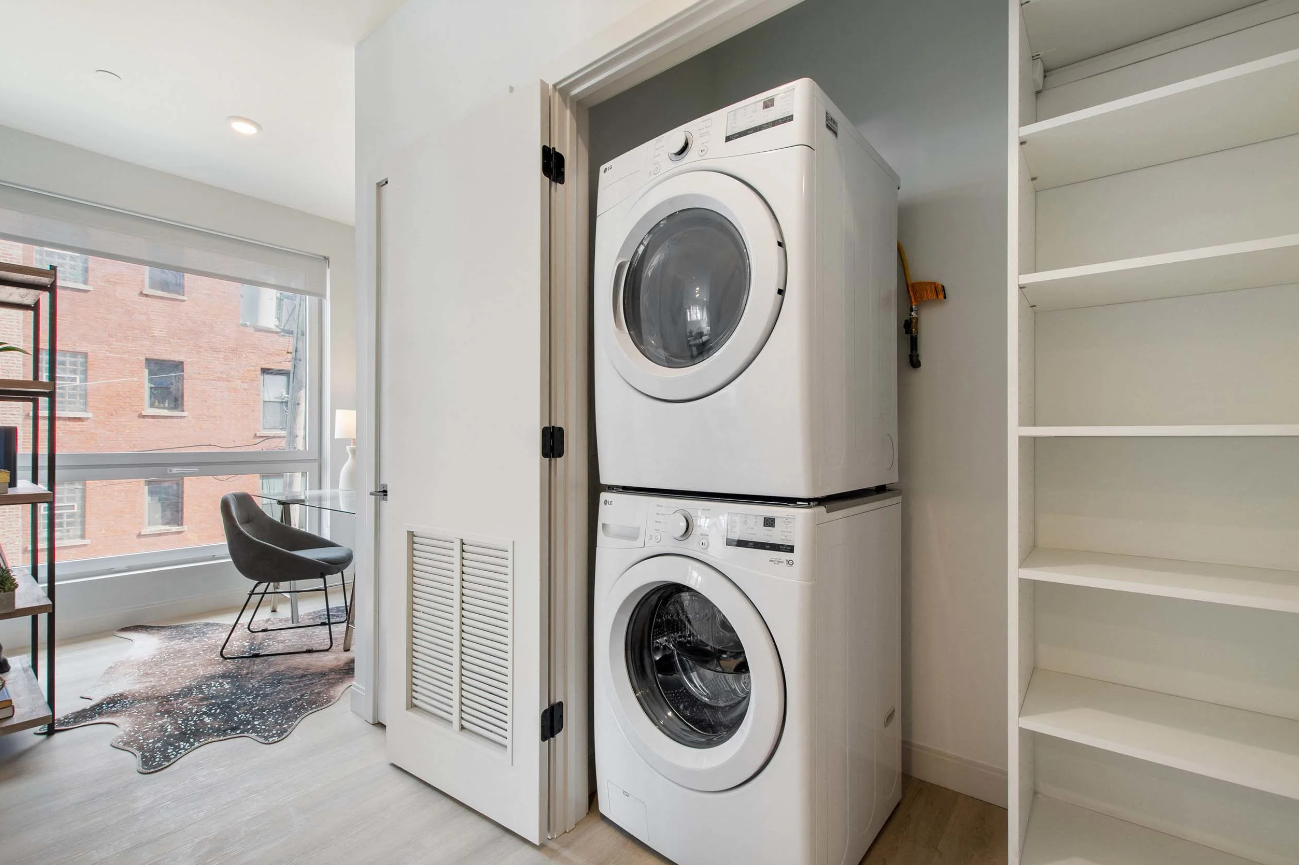 Stacked washer and dryer in a bright closet beside open shelves and a windowed room.