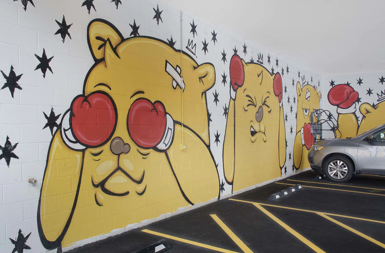 Cartoon bear mural with red heart-shaped sunglasses on a wall beside a parked car in a lot