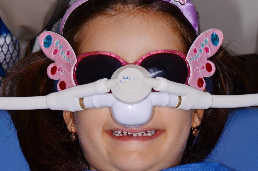 Bambina sorridente con occhiali da sole rosa a forma di farfalla e una sorta di mascherina bianca su naso e bocca.
