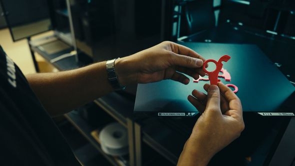 Person holding a red, 3D-printed object in front of a 3D printer.