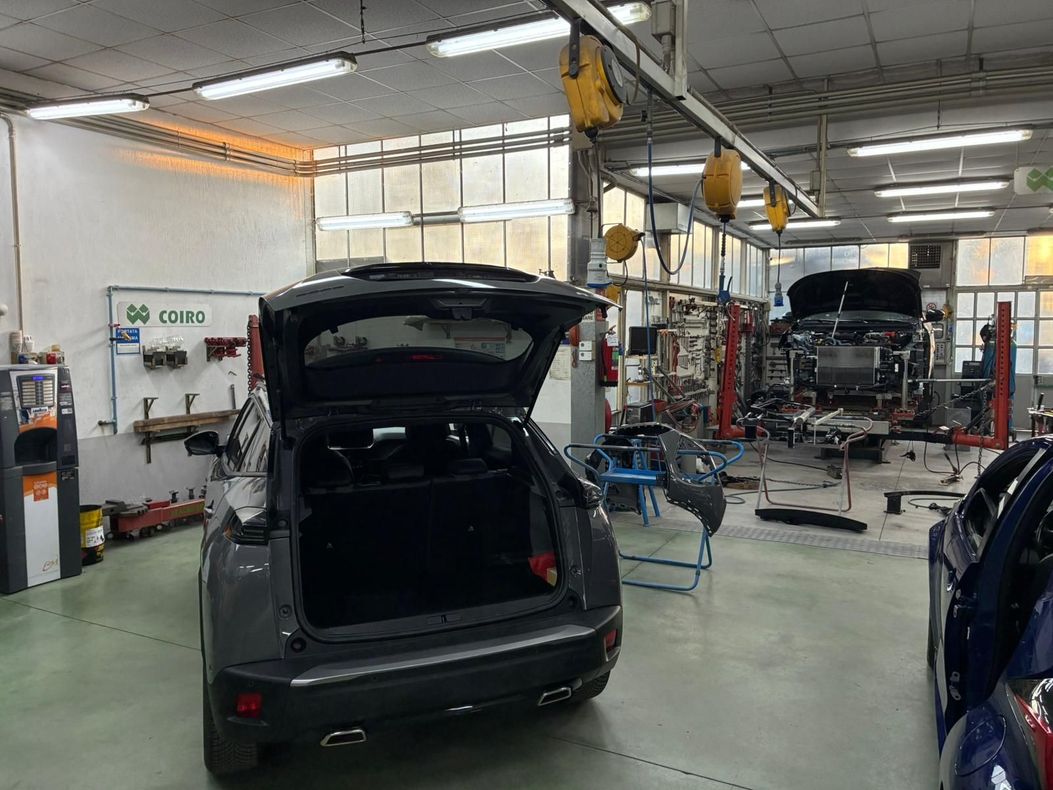 Auto con bagagliaio aperto in officina, un'altra auto sul ponte sollevatore sullo sfondo. Sono visibili gli attrezzi e le attrezzature dell'officina.