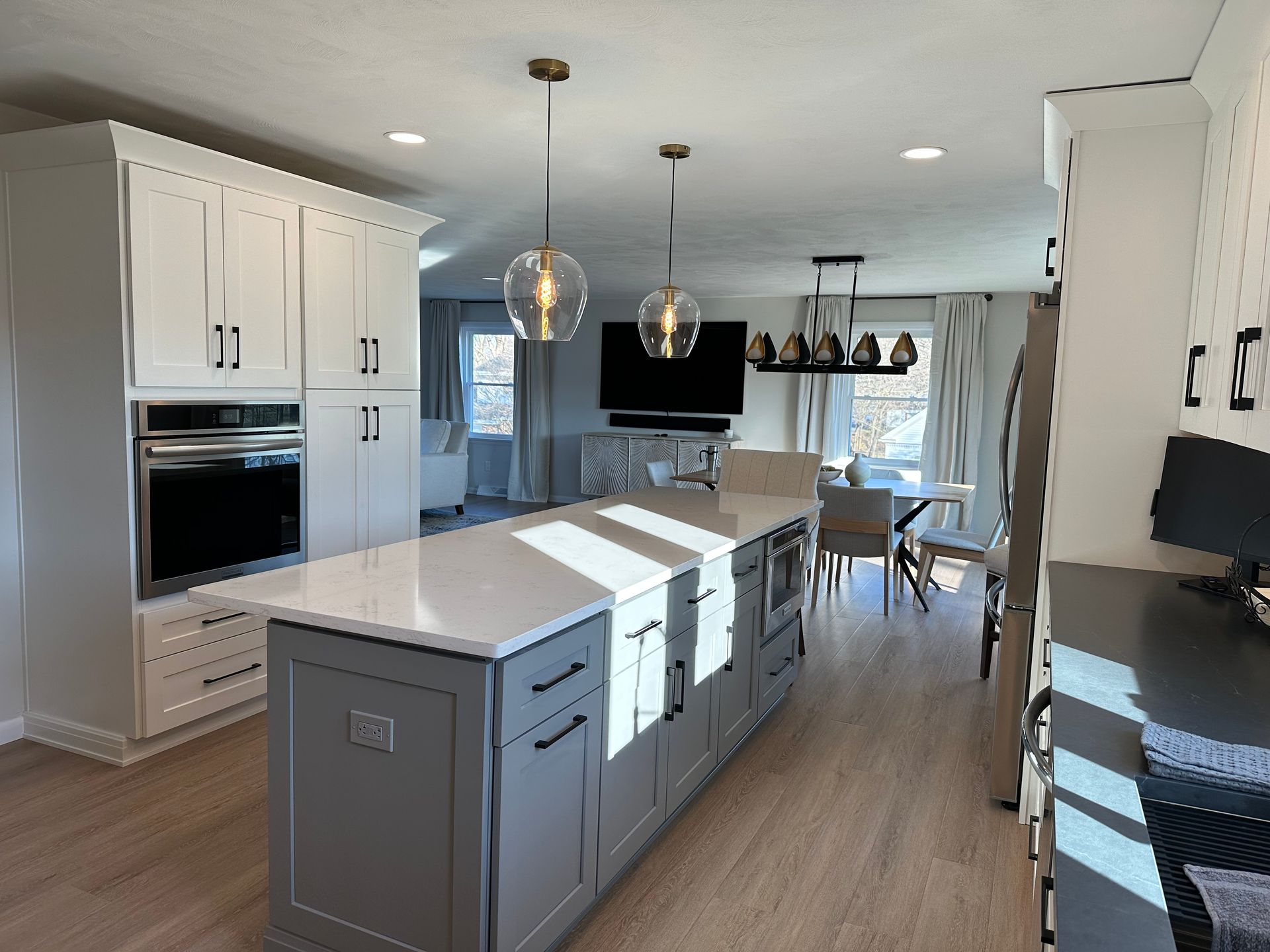 Modern kitchen with gray island, white cabinets, and two pendant lights. Dining area and TV in the background.
