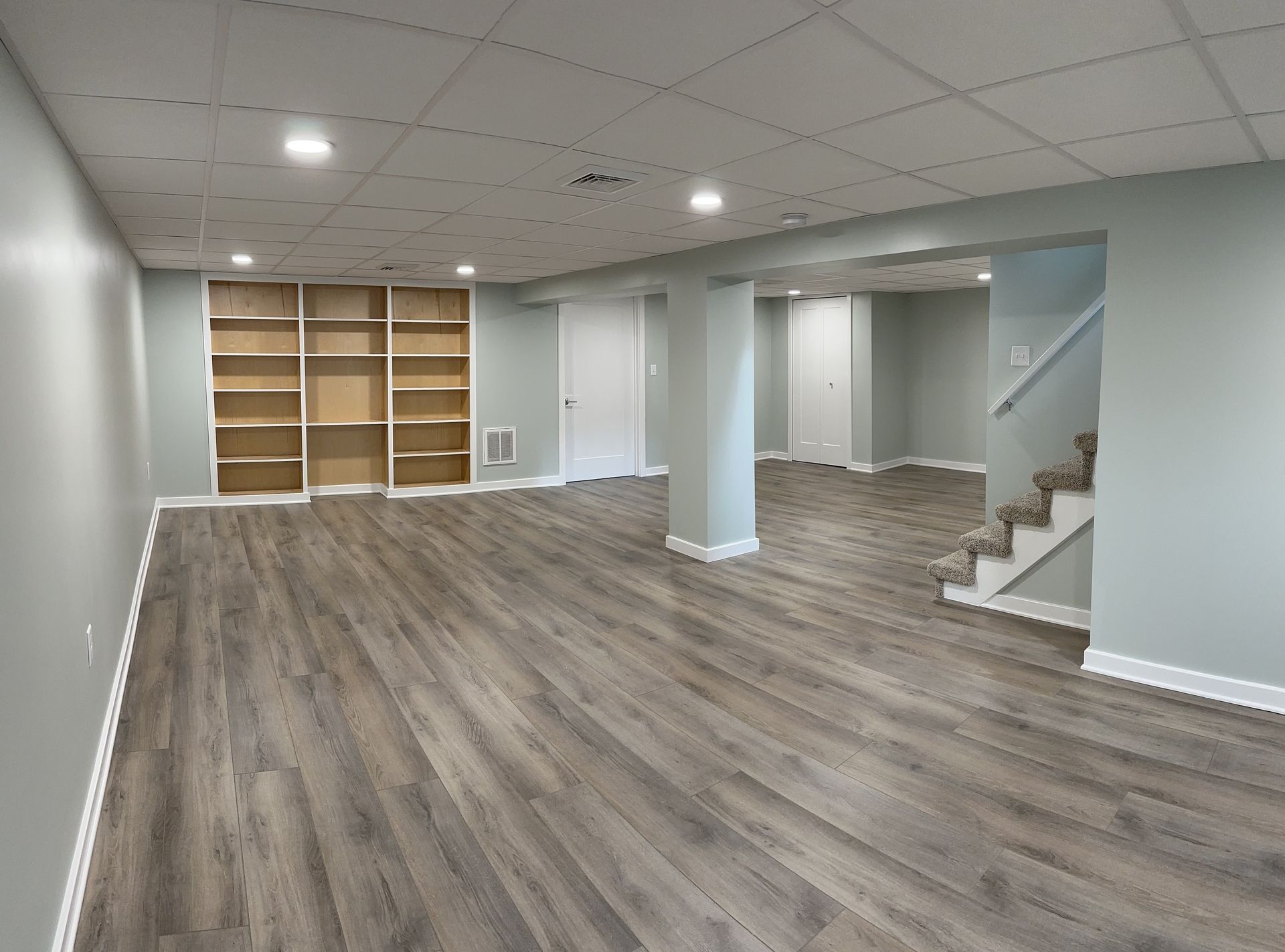 Empty, newly renovated basement with light gray walls and wood-look flooring. Includes built-in shelving and a staircase.