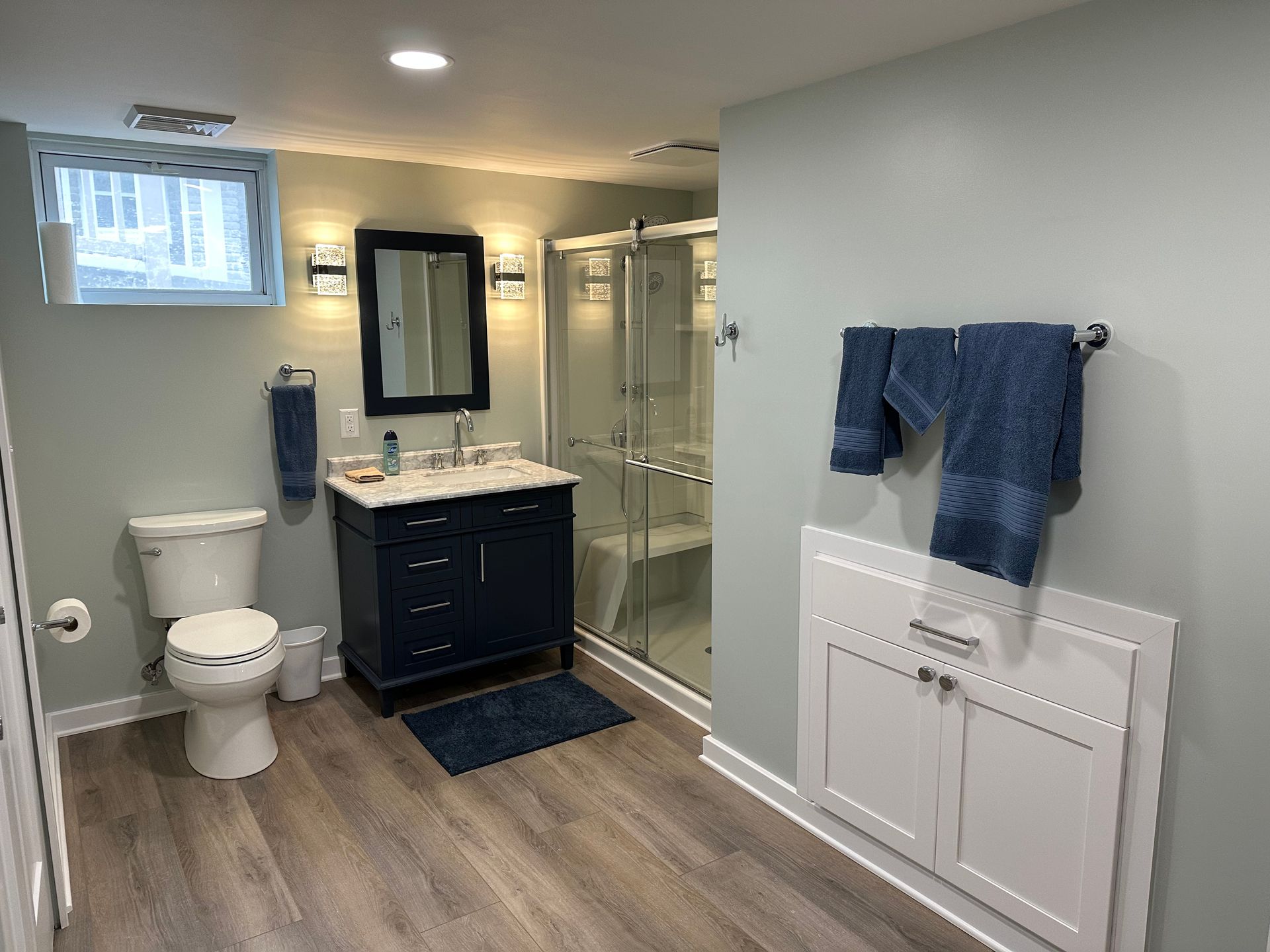 Bathroom with blue walls, navy vanity, glass shower, toilet, and folded cabinet. Wooden flooring.