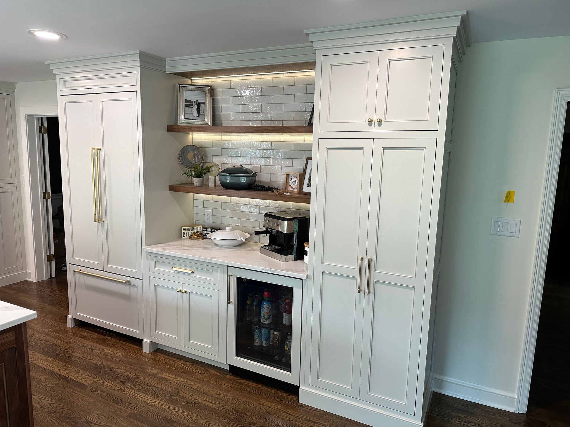 White built-in kitchen cabinets with refrigerator, coffee station, and wine cooler. Wood flooring.