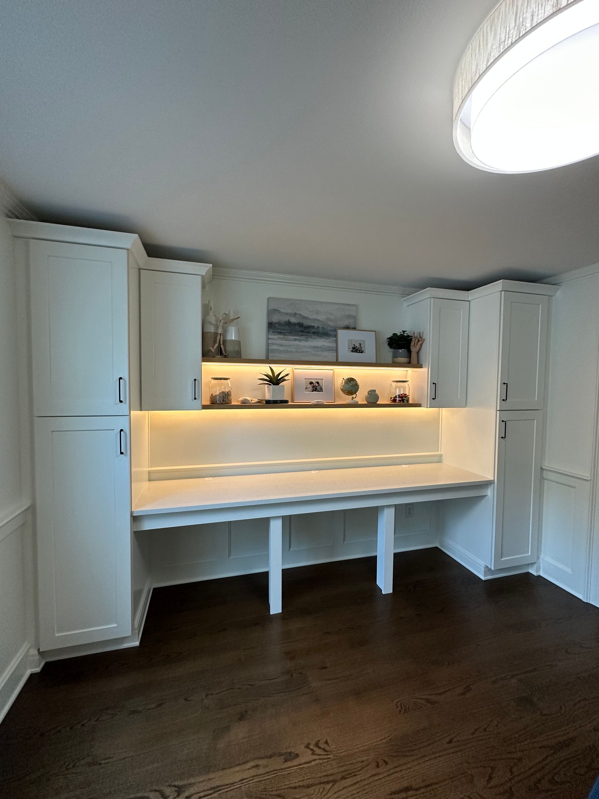 Built-in white desk with storage cabinets and a lighted shelf against a white wall in a room with dark wood floors.