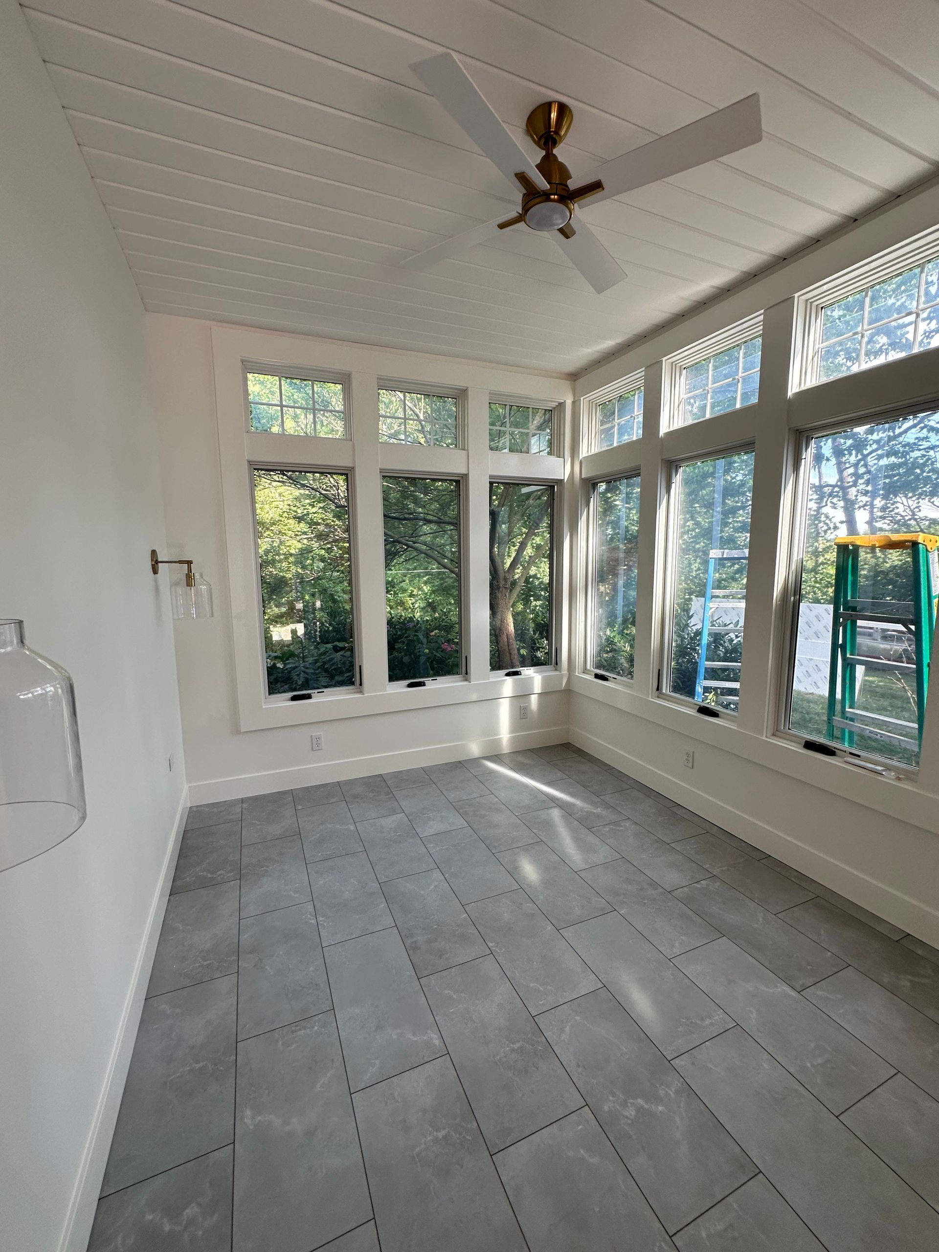 Sunroom with grey tiled floor, many windows, white walls, and ceiling fan. Exterior view of trees.